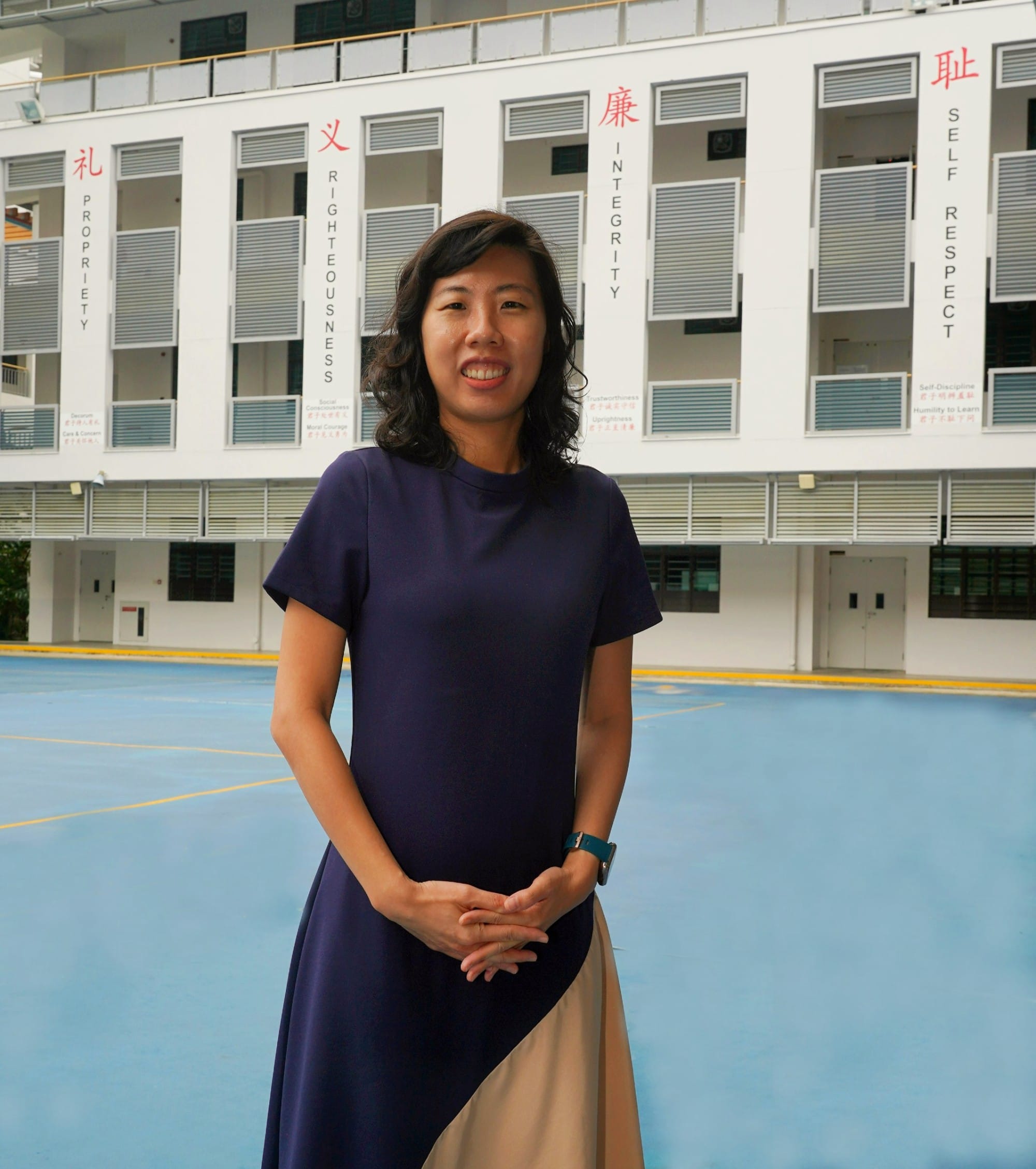 Woman in navy dress stands on a blue school court, with a building showing values like Integrity and Righteousness behind her.